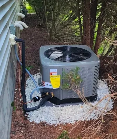 Gray AC condenser unit installed on gravel pad with refrigerant lines and electrical connections